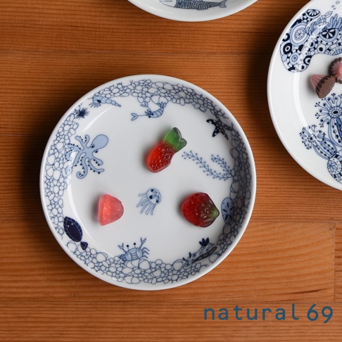 白色陶瓷小碟，飾有藍色海洋生物圖案，包括章魚、水母、螃蟹和魚。碟上擺放著三顆造型為草莓的軟糖，顏色為紅色和綠色。