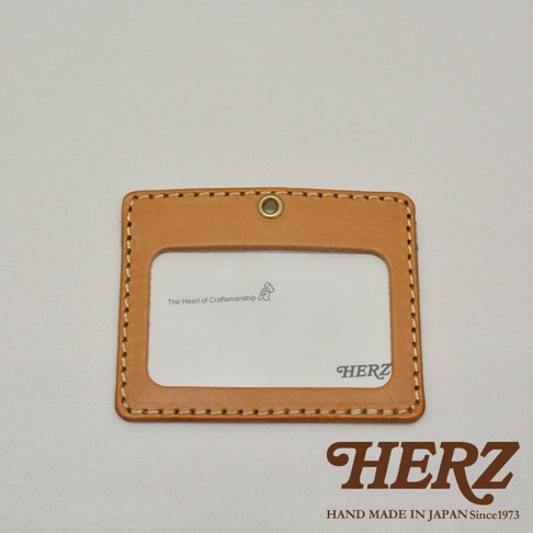 職人手作皮革橫式證件識別夾，淺棕色皮革，車縫線明顯，正面有透明視窗，上方有金屬掛孔，印有「The Heart of Craftsmanship」及小圖標，右下角印有「HERZ」字樣。