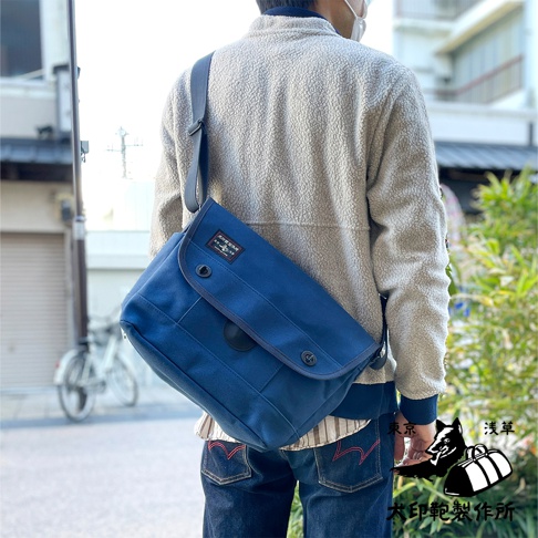 一個藍色的純棉帆布雙扣郵差包，由一名男性背著，包包上有一個黑色皮革標籤，包包的翻蓋上有兩個金屬扣。