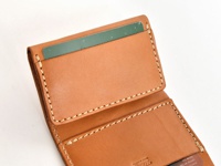 職人手作皮革三折式迷你短夾，採用淺棕色皮革，內裡有卡槽，卡夾內插著一張綠色卡片，車縫線為米白色。