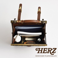 職人手作皮革復古口金後背包，棕色皮革材質，內部裝有文件夾、筆記本、小錢包和圓形白色小物。背包採用金屬口金開合，配有兩條可調節的皮革揹帶和提把。