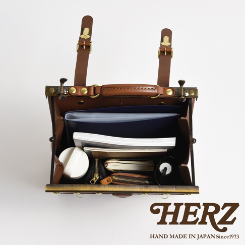 職人手作皮革復古口金後背包，棕色皮革材質，內部裝有文件夾、筆記本、小錢包和圓形白色小物。背包採用金屬口金開合，配有兩條可調節的皮革揹帶和提把。