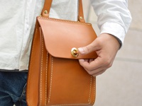 焦糖色厚實皮革單肩包，帶有金色旋轉鎖扣和白色縫線細節，由手持展示。