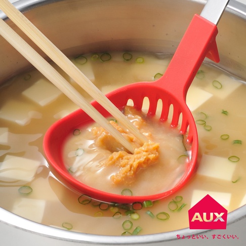 紅色不沾桌多用濾水湯匙，包含湯匙頭的側邊濾孔，用兩根木質筷子夾著味噌，在裝有豆腐和蔥花的湯鍋中操作。