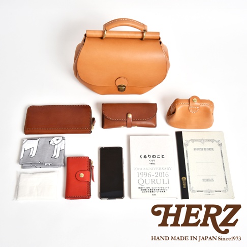 一張從上方俯視的照片，展示日本職人品牌HERZ的手作皮革包與隨身物品。畫面中央為一個淺棕色皮革屋瓦風包款，周圍擺放了長錢包、小型皮革收納包、紅色鑰匙包、繪有小狗圖案的布巾、衛生紙包、智慧型手機以及一本名為Kuruli 20th Anniversary的紀念書籍和一本Herz筆記本，右下角印有Herz品牌標誌與手工製作字樣。