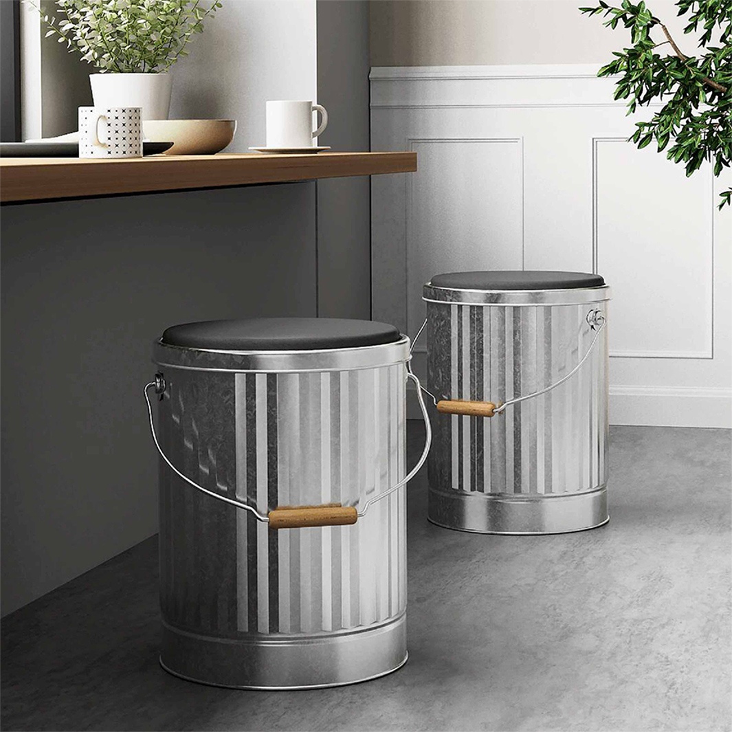 兩個銀色金屬桶凳，頂部配有黑色坐墊，其中一個放在另一個前面，後者靠在白色的牆壁裝飾旁邊。