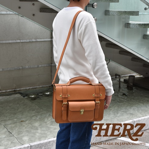 職人手作皮革學院風兩用商務公事包，採用天然皮革製成，呈現溫潤的焦糖棕色。包款設計簡潔，配有手提把、可調式長背帶、雙皮帶釦開合，以及前方小口袋，適合商務或日常使用。