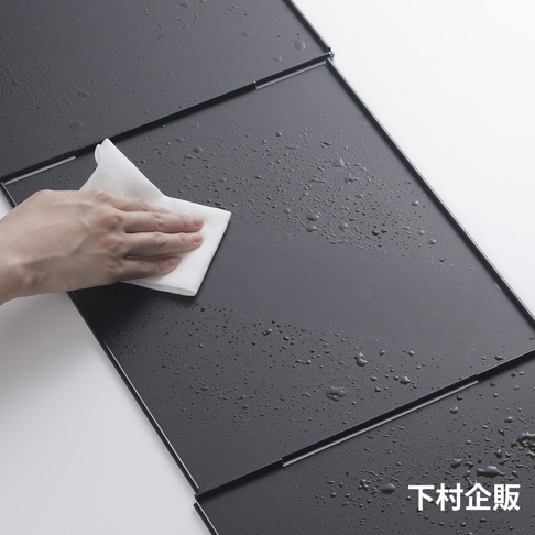 深灰色ㄇ型折疊防汙擋油板，表面有水珠，一隻手正用白色抹布擦拭。