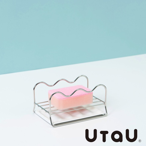 不鏽鋼製的萬用瀝水支架，上面放置著粉紅色海綿，支架設計簡約，邊緣呈波浪狀，適合置於廚房或浴室檯面。