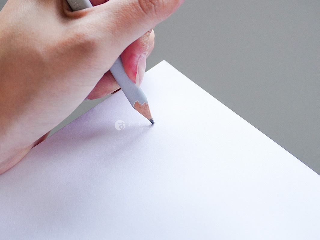 手持淺灰色鉛筆，在純白色紙張上書寫。紙張邊緣呈現柔和的淡紫色光澤，背景為淺灰色。