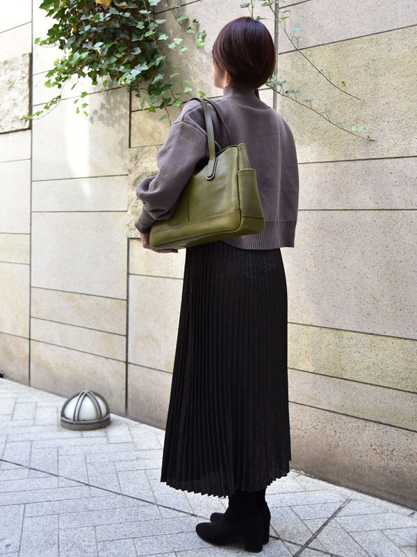 一位女士身著深灰色高領毛衣和黑色百褶長裙，肩上挎著一個橄欖綠色皮革橫式肩背托特包。