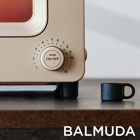 The Toaster 蒸氣烤麵包機，奶茶限定色，機身為淺米色，設有圓形旋鈕，標示著時間刻度和「ON/OFF」字樣。旁邊有一個黑色的小咖啡杯。