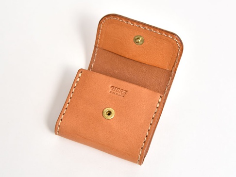 一個淺棕色皮革製成的雙折收納零錢包，帶有黃銅按扣和縫線細節，標有「HERZ MADE IN JAPAN」字樣。