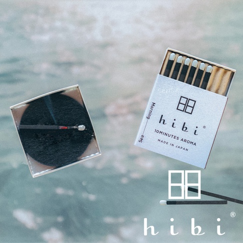 此為 hibi 香氛火柴禮盒，右側盒子上方展示一排未點燃的黑色火柴，盒身印有 hibi 品牌標誌與 10 MINUTES AROMA 字樣。左側放置一個黑色圓形香氛墊，上面橫放著一根正在燃燒的火柴。右下角則有品牌商標與兩根火柴的圖案，整體呈現簡約日式風格。