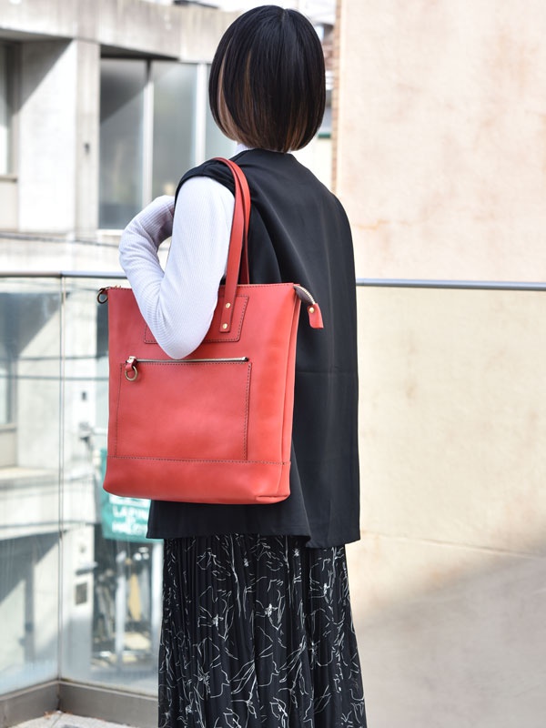 一位女士提著一個紅色皮革直式拉鍊托特包，包包上有一個外部拉鍊口袋和一個金屬環。