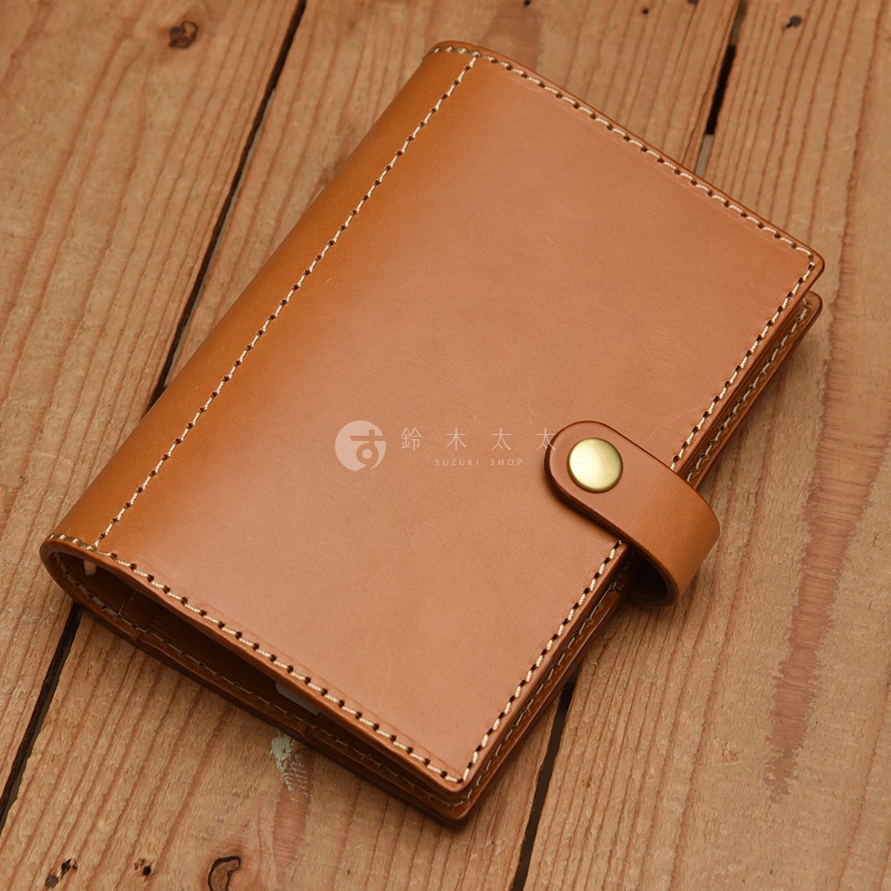 淺棕色皮革手帳，職人手作，以白色縫線勾勒邊緣，配有金色按扣封口，放置於木紋桌面。