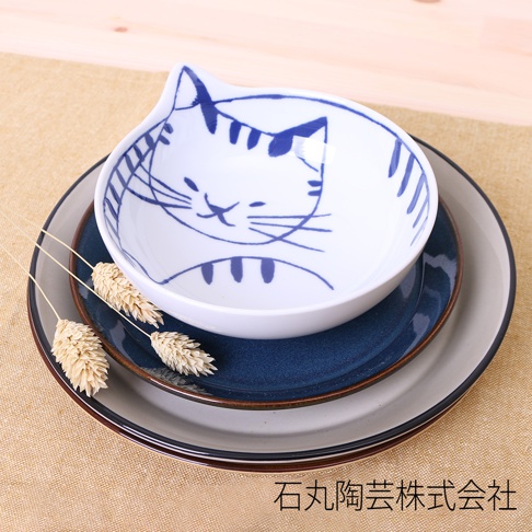 白色陶瓷碗，碗內繪有藍色線條的貓咪臉，碗邊呈貓耳狀。碗下方疊有兩個不同尺寸的深藍色和灰色盤子，盤子邊緣有細微的棕色線條。三支乾燥的稻穗置於碗旁。