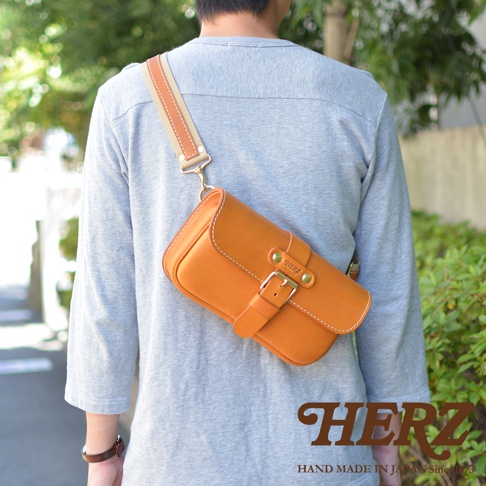 一名男子背著一個亮棕色皮革單扣斜背包，包包上有白線縫邊、金色金屬扣和品牌標誌。