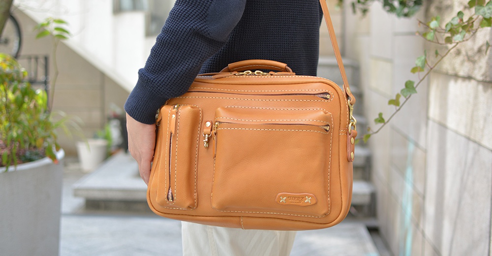 職人手作的淺棕色皮革兩用單肩包，有多個拉鍊口袋和一個翻蓋口袋，包身上有縫線細節，並帶有品牌皮革標誌。