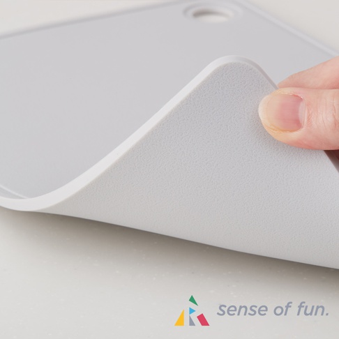 淺灰色矽膠砧板，材質柔軟，邊緣圓潤。一隻手正在彎曲砧板的一角，展示其彈性。砧板表面帶有細微紋理，右上角有一個圓形掛孔。