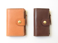 兩個職人手作皮革拉鍊零錢鑰匙包，一個為淺棕色，一個為深棕色，皆配有金色拉鍊及同色系皮革拉繩，並以撞色縫線勾勒出包身。