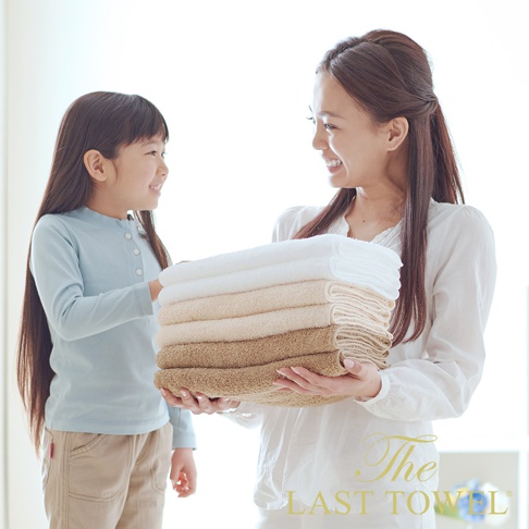 堆疊的純棉長毛巾，呈現白色、米色和淺棕色漸層。一位女士雙手捧著毛巾，一位小女孩站在旁邊。