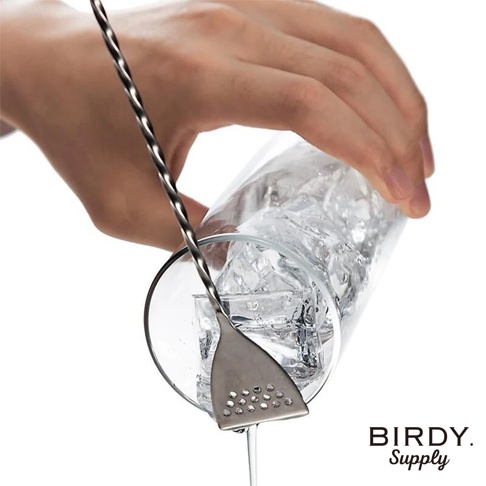 手持一支30公分長的不鏽鋼調酒攪拌匙，其螺旋紋路狀的長柄末端連接一個扁平的鏟狀頭，鏟頭上有規則排列的小孔，正插入裝有冰塊與水的玻璃杯中。