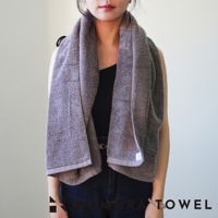 一個穿著黑色上衣和牛仔褲的女人，身上圍著一條灰褐色的毛巾，毛巾上有 NOMURA TOWEL 的字樣。