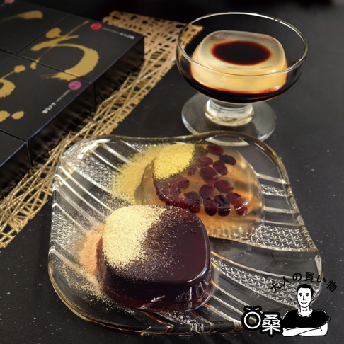 精緻的透明玻璃盤上擺放著兩塊呈現凍狀的和菓子，一塊為深褐色並撒有黃豆粉，另一塊為透亮的琥珀色且內嵌紅豆。盤後方配有一杯裝盛在玻璃腳杯中的甜點，上方淋有深色糖漿，左側則有印著金色書法字樣的黑色禮盒，整體畫面呈現質感優雅的日式甜點擺設。