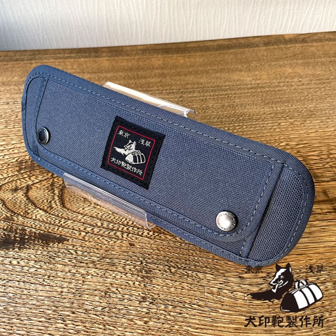 深藍色純棉帆布製成的長方形收納包，帶有銀色按扣和一個黑色方形刺繡布標，上面印有狗的圖案和文字。