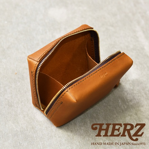一個 tan 色皮革製的箱型收納包，有金色拉鍊，包包內有隔層，品牌標誌 Herz 壓印於底部。