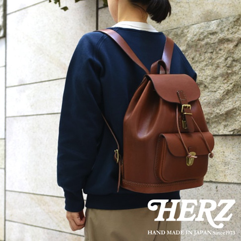 棕色皮革後背包，採用經典束口設計，配有翻蓋和金屬鎖扣，側邊有方形口袋，肩帶為皮革材質，整體呈現復古風格。