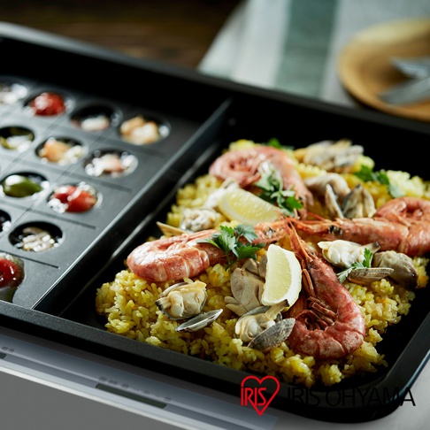 黑色電烤盤分為兩區，左區為圓形烤模，盛有各式小食；右區鋪滿黃色米飯，上面擺放著鮮紅的蝦子、蛤蜊與檸檬片，旁邊有綠色香草裝飾。