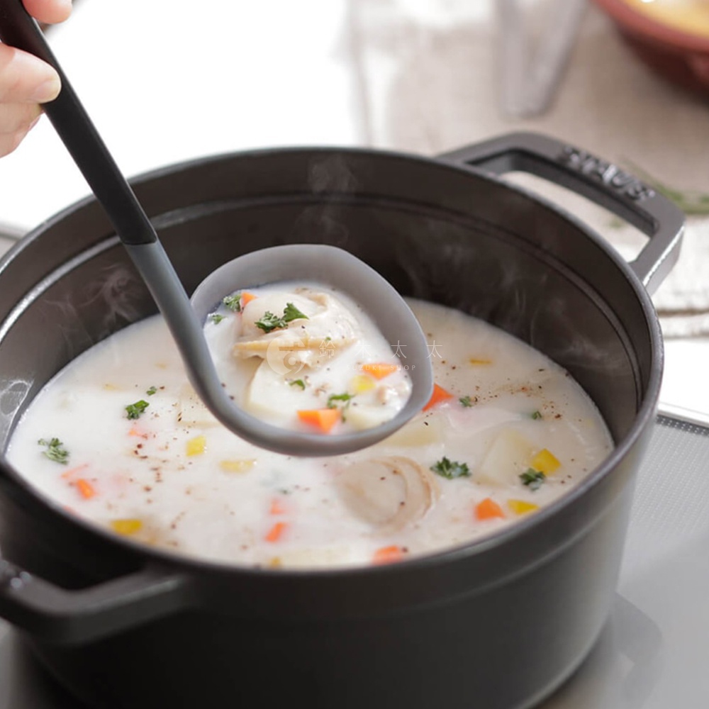 一個手持灰黑色矽膠湯勺，正在從黑色湯鍋中舀起奶油濃湯，湯裡有塊狀的馬鈴薯、紅蘿蔔丁和香草。