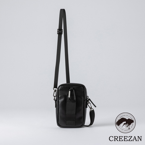 黑色皮革迷你側背收納包，表面有顆粒紋理，配有可調節的黑色尼龍斜背帶，包身正面有黑色織帶及登山扣裝飾。