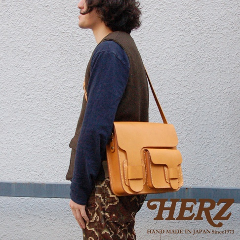 淺棕色皮革製成的厚實單肩包，包身有兩個翻蓋口袋，縫線明顯，由日本品牌 HERZ 製造。