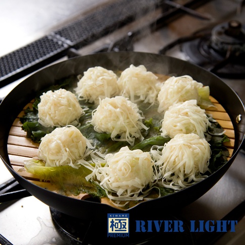 黑色的 30 公分炒鍋內，擺放著竹蒸籠，蒸籠上層層疊疊著數個白色、外觀類似燒賣的食物，食物表面覆蓋著細絲狀的配料，下方襯著綠葉菜，鍋中冒著蒸氣。