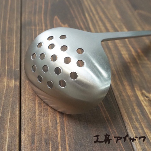 不鏽鋼萬用瀝水湯勺，勺頭為橢圓形，佈滿圓形瀝水孔，勺柄為長條形，整體呈現銀色金屬光澤，置於木紋桌面。