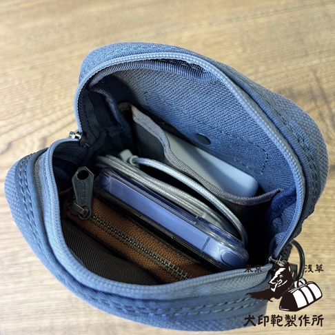 一個打開的深藍色帆布手機斜背包，內部設有分層收納空間，放置了手機、充電線與小型錢包。畫面右下角帶有犬印鞄製作所的品牌圖示與文字，整體呈現簡潔實用的收納細節。