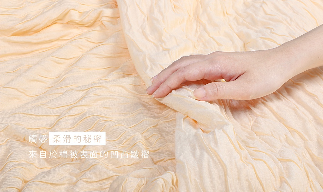淺黃色舒芙蕾透氣涼被，無棉款，織紋細膩，呈現柔軟的褶皺質感，一隻手輕柔地撫摸著涼被表面。
