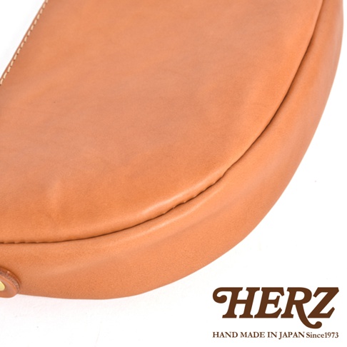 日本 HERZ 品牌皮革半圓馬鞍包特寫，淺棕色皮革質感，細膩縫線，底部有品牌 LOGO。
