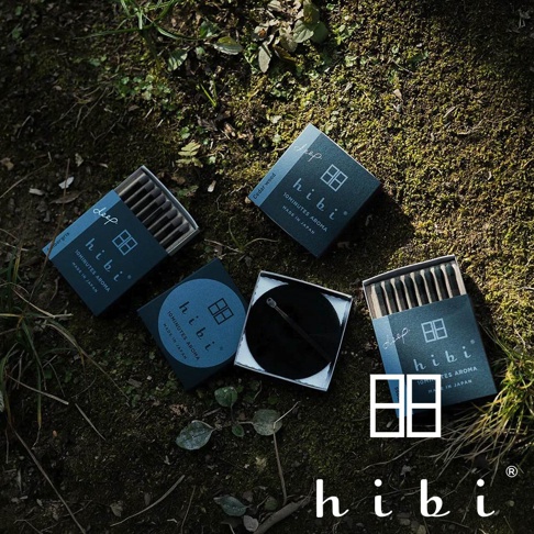 俯拍視角呈現的三盒 hibi Deep 系列木質調香氛火柴，外盒為沈穩的深色設計，旁邊擺放著一個圓形專用燃燒墊，所有商品皆散落在帶有綠色苔蘚的自然泥土地上，展現出品牌日系簡約與森林氣息的產品風格。