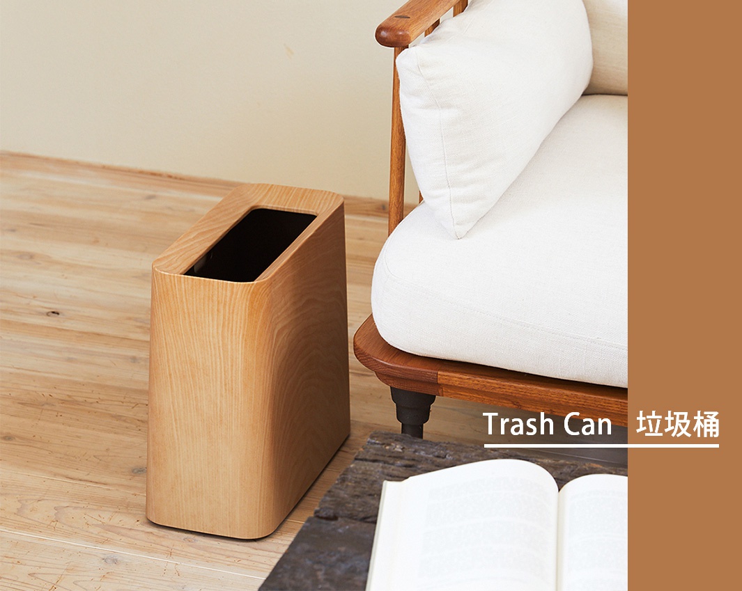 一個淺木色、帶有橡木紋理的方形垃圾桶，頂部開口為長方形，側面圓潤，放置在木質地板上，旁邊是一張帶有白色坐墊的木質椅子。