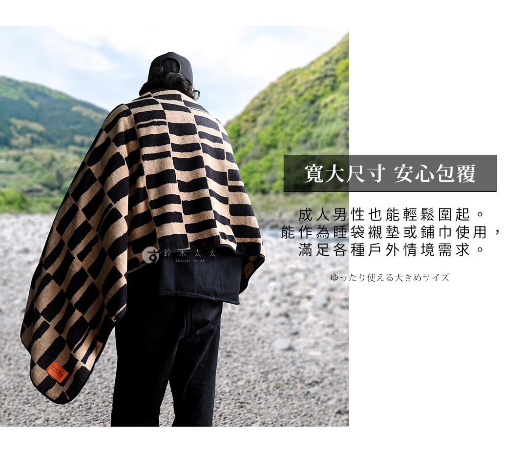 從背後拍攝，一位穿著黑色帽 T 和牛仔褲的人，披著一條由黑色和米色格紋組成的寬大毛巾。毛巾材質看起來柔軟，上面有日文和英文的品牌標誌。背景是模糊的山景和河邊。