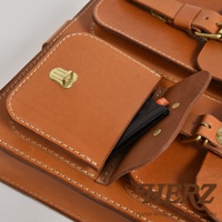 職人手作皮革三口袋後背包，採用天然皮革製作，呈現溫潤的焦糖棕色。包款設有三個外口袋，其中一個口袋露出部分黑色物品，上面印有橘色JBL標誌。包款以手工縫線加固，搭配黃銅色金屬鎖扣和調節扣。