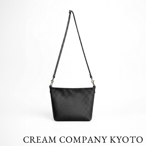 一個由日本京都品牌 CREAM COMPANY 製造的黑色皮革側背包，採用質感細膩的荔枝紋牛皮製成，設計簡約輕巧，配有可拆卸的細肩帶，包身中央下方標示著 CREAM COMPANY KYOTO 的品牌字樣，背景為純白。