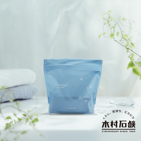 淺藍色帶有拉鍊封口的洗衣粉包裝袋，袋上印有白色字體「LAUNDRY POWDER」及「asuno」字樣。背景有疊放的白色與灰色毛巾，以及綠色植物葉片。