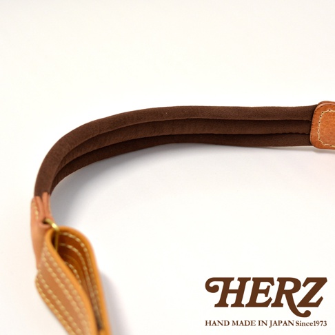 職人手作皮革鹿角扣繩郵差包的特寫，展示了咖啡色麂皮襯裡的肩帶，以及 tan 色皮革與縫線的細節。