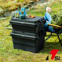 兩個黑色塑膠可堆疊收納箱，帶有銀色金屬扣環。箱子頂部擺放有藍色水壺、露營瓦斯罐、油燈和一把橘色小刀。背景為草地和模糊的樹木。