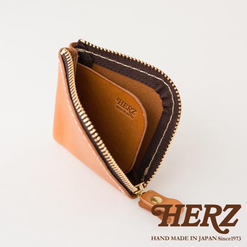 一個棕色皮革材質的日本HERZ職人手作零錢包，採用L型拉鍊設計，內部設有皮革隔層，整體呈現簡約精緻質感，右下方標有品牌標誌與日本製造字樣。